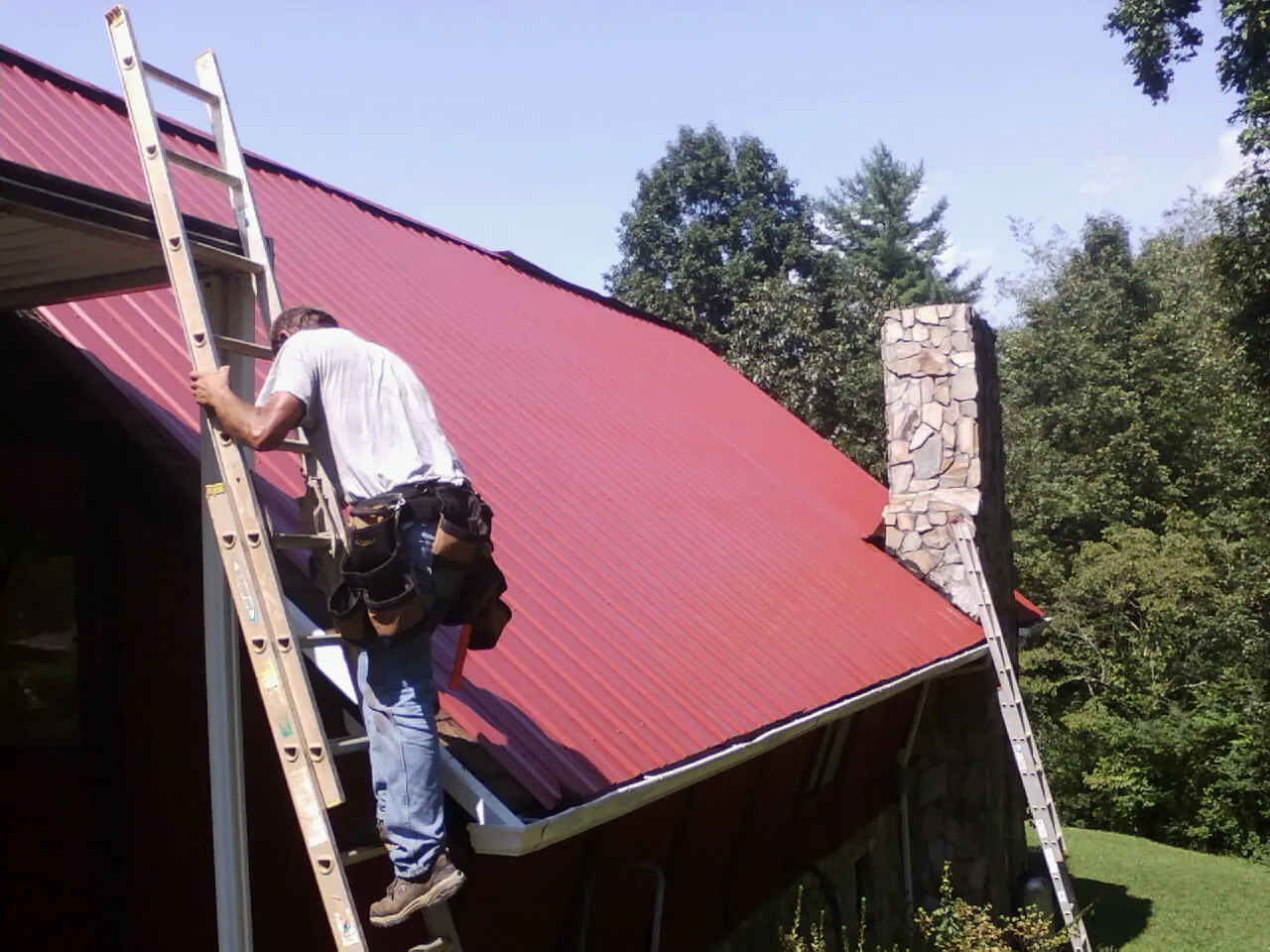 Roofer on a ladder beside a red metal roof and stone chimney — Soffit & Fascia Repair by Roofing Brothers in Rock Falls